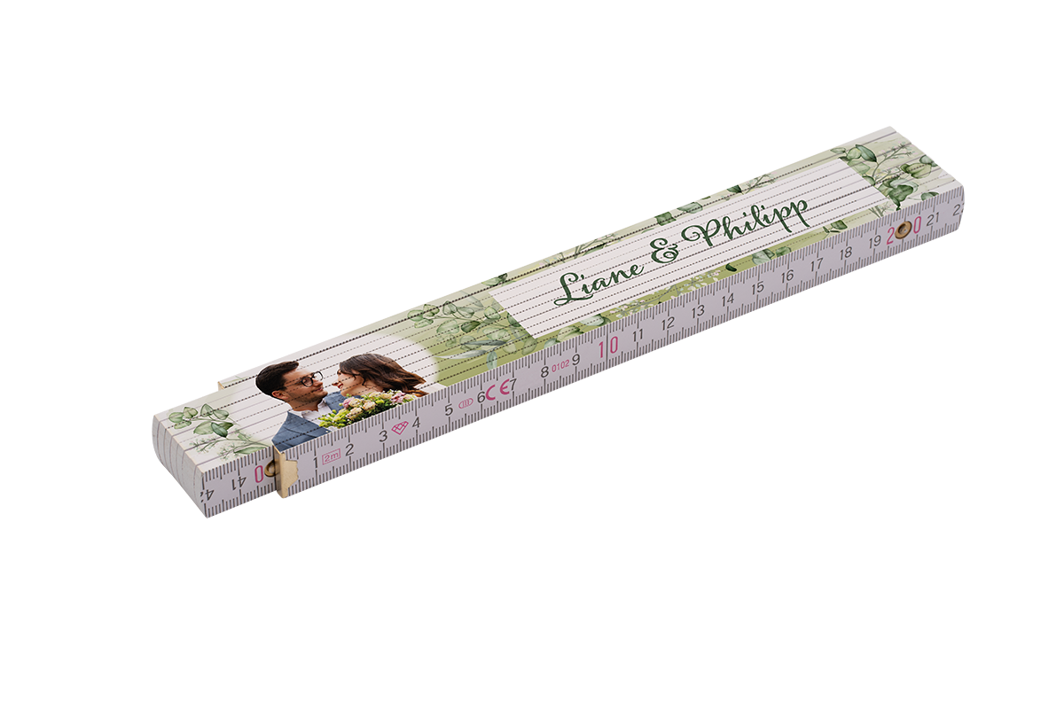 Gefalteter weiß bedruckter Zollstock mit grünen Eukalyptus-Illustrationen, aufgedrucktem Foto eines Paares mit Blumenstrauß, kursiver Aufschrift „Liane & Philipp“, sichtbaren Zentimeter-Markierungen und Messskala sowie Messinggelenken.