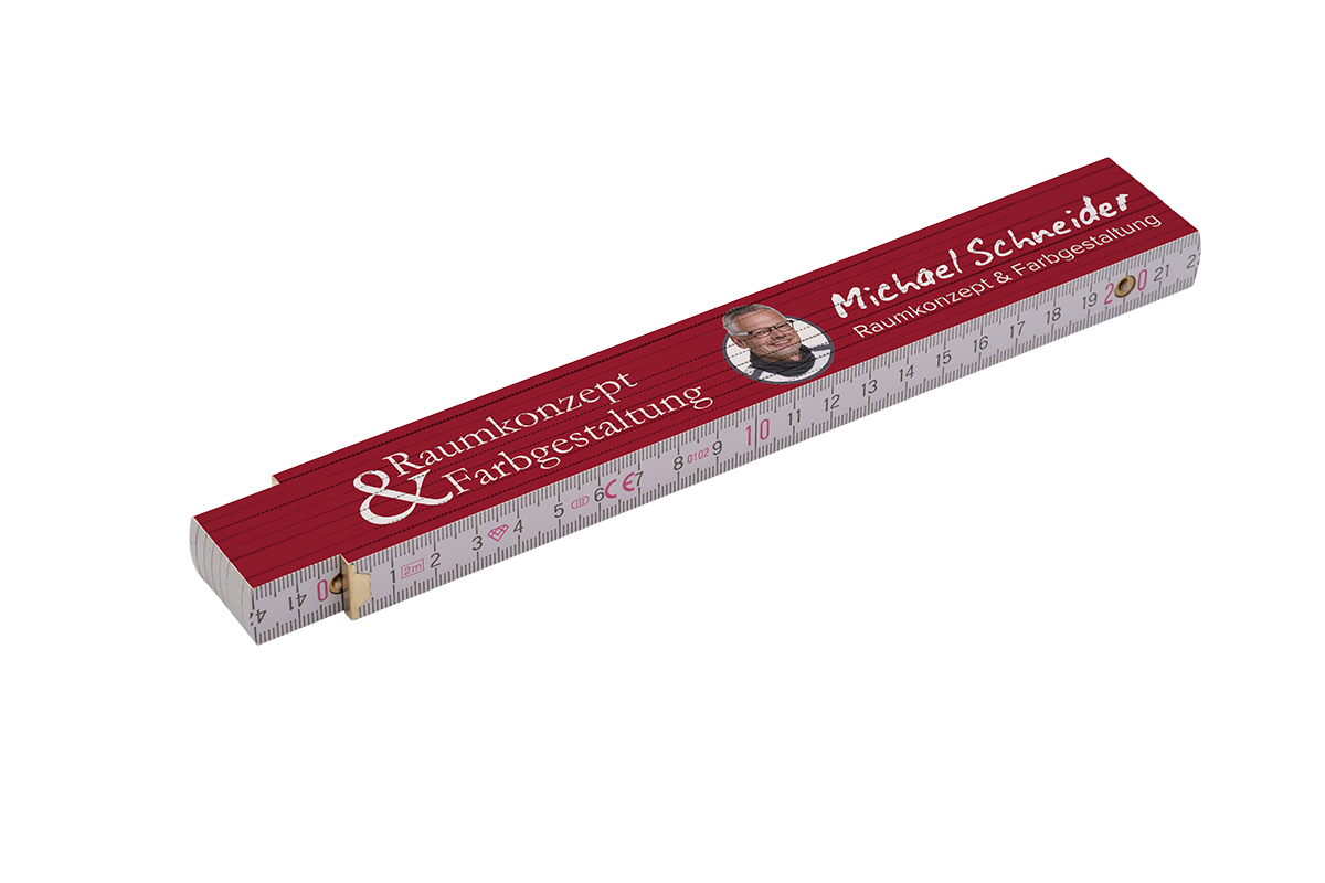 Klappmeter mit dunkelroter Deckfläche, aufgedrucktem Schriftzug „Michael Schneider – Raumkonzept & Farbgestaltung“, sichtbarer Zentimeter-Skala und kleinem rundem Porträt.