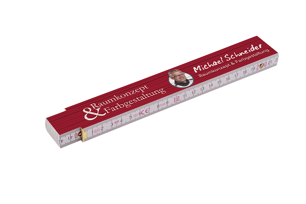 Roter zusammengeklappter Holz-Zollstock mit sichtbarer Zentimeter-Skala, auf der Oberseite gedrucktem Schriftzug Michael Schneider und Raumkonzept & Farbgestaltung sowie einem kleinen runden Porträtfoto eines lächelnden Mannes.